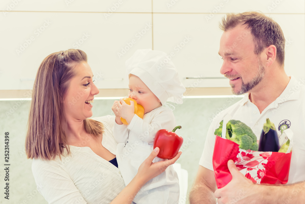 Happy parents standing in the kitchen and prepares for cooking with their baby girl chef. Father bring vegetables in paper bag. Baby girl made a grimace. Happy parents standing in the kitchen and prepares for cooking with their baby girl chef. Father bring vegetables in paper bag. Baby girl made a grimace.