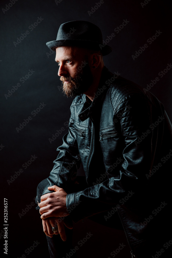 Cool hipster in hat and leather jacket looking away on black background Cool hipster in hat and leather jacket looking away on black background