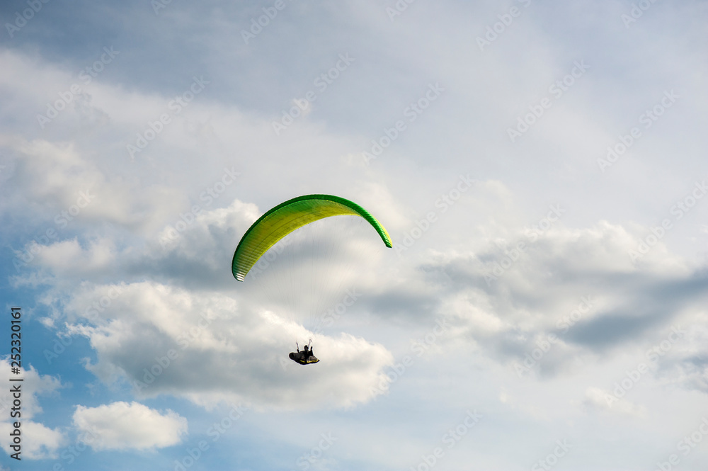 Yellow and green paraglider is flying in the blue sky against the background of clouds. Paragliding in the sky on a sunny day. Yellow and green paraglider is flying in the blue sky against the background of clouds. Paragliding in the sky on a sunny day.