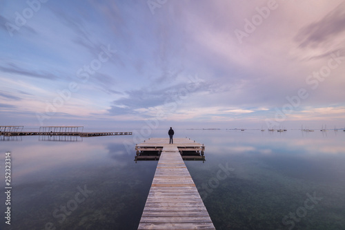 Back view of unrecognizable person standing on long lumber pier near calm sea water on cloudy evening in Spain photo