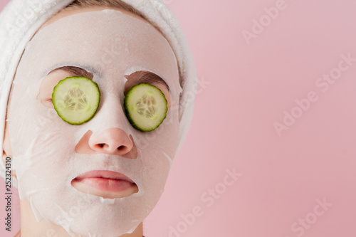 Beautiful young woman is applying a cosmetic tissue mask on a face with cucumber on a pink background