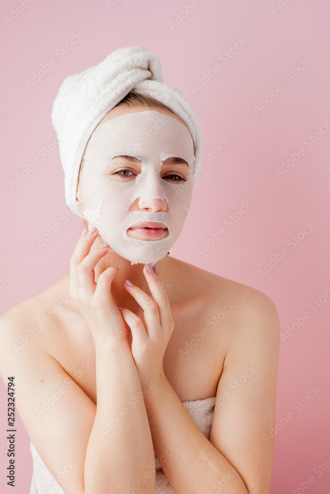 Beautiful young woman is applying a cosmetic tissue mask on a face on a pink background. Healthcare and beauty treatment and technology concept Beautiful young woman is applying a cosmetic tissue mask on a face on a pink background. Healthcare and beauty treatment and technology concept