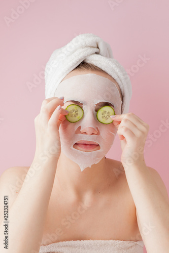Beautiful young woman is applying a cosmetic tissue mask on a face with cucumber on a pink background. Healthcare and beauty treatment and technology concept