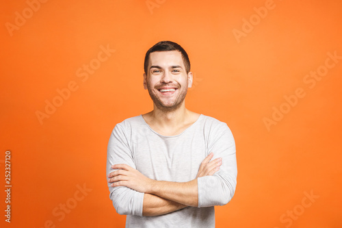 Casually handsome. Confident young handsome man keeping arms crossed and smiling while standing against orange background.