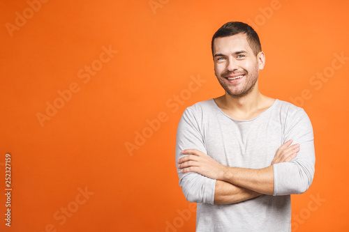 Casually handsome. Confident young handsome man keeping arms crossed and smiling while standing against orange background.