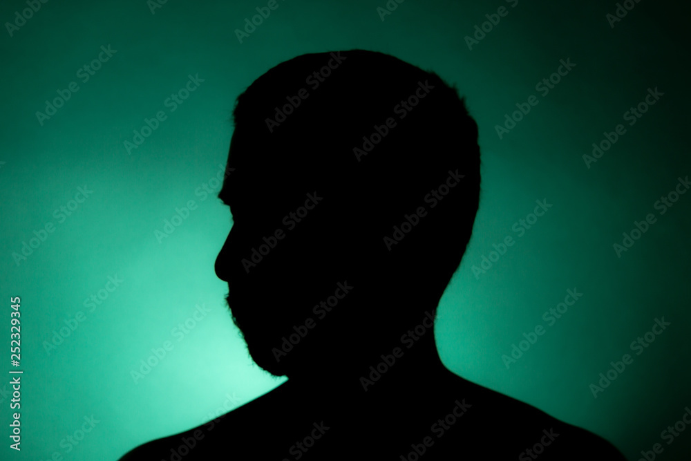 Cleanly defined silhouette of a male person turned to the left against a green background with a spotlight and bright area right behind the bust. Studio shot with well defined colour background.