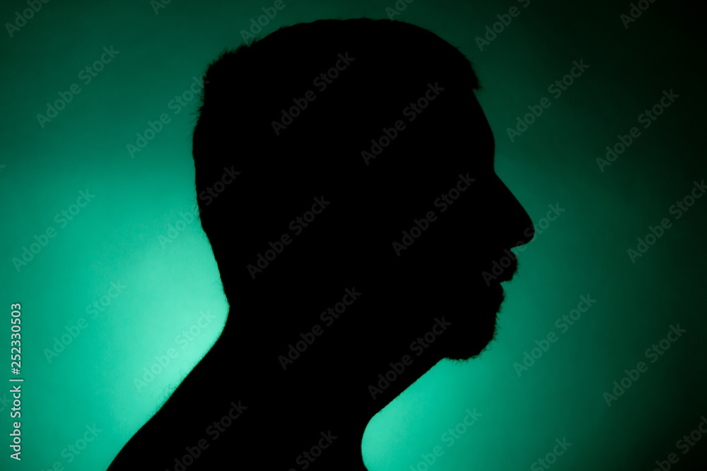 Cleanly defined silhouette of a male person turned to the left against a green background with a spotlight and bright area right behind the bust. Studio shot with strong explicit colour background.