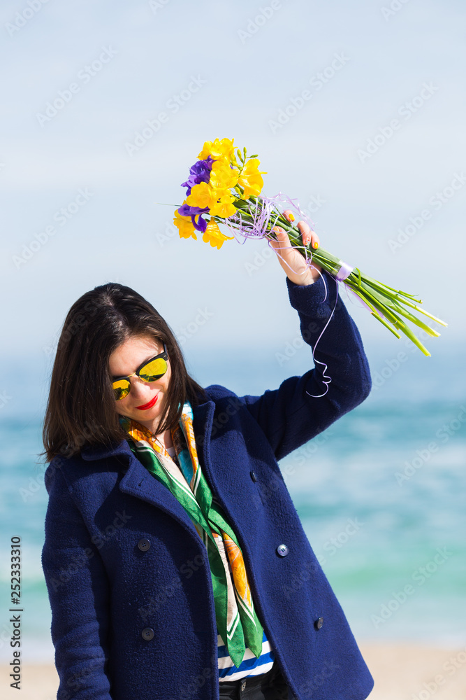 Φωτογραφία Stylish young woman in sunglasses and coat holding bouquet of freesias and irises with sea landscape on blurred background