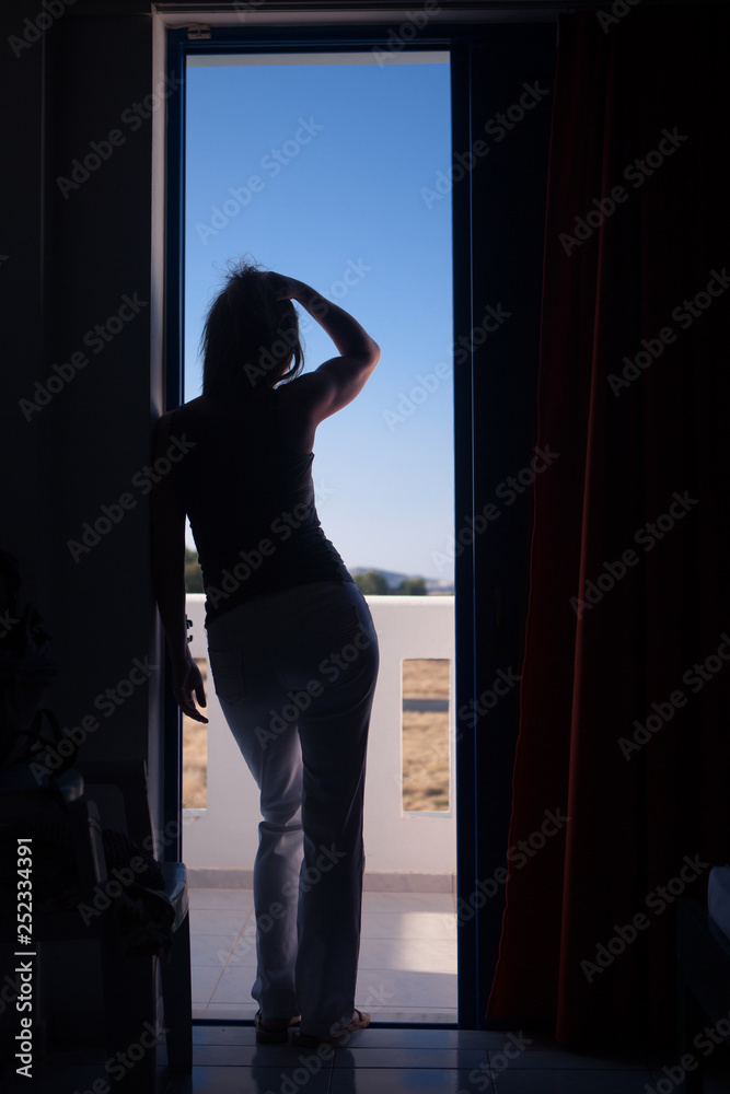 Lockdown, Stay home. Sporty woman stands silhouetted in the balcony door and runs her hand through her hair. She looks into a mediterranean landscape.
