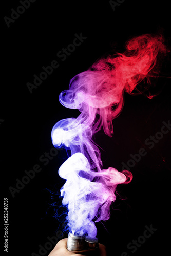 Male hand holds vape. Vape clouds splash from vape coil on black background. Fog is dual color     blue and red. Stock isolated dual colour smoke with spray boiling glycerine.