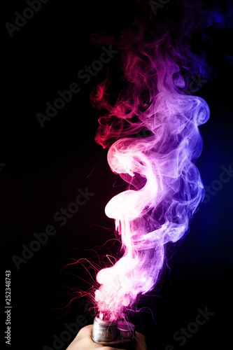 Fototapeta Naklejka Na Ścianę i Meble -  Male hand holds vape. Vape clouds splash from vape coil on black background. Fog is dual color — blue and red. Stock isolated dual colour smoke with spray boiling glycerine.