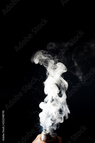 Man hand holds vape. Vape clouds splash from coil on black background. Fog is white. Stock isolated white smoke with spray boiling glycerine around. © Alexs