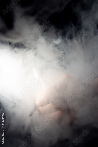 Male hand holds vape in thick vape fog or smoke. Vape clouds around hand with vape on black background. Fog is white. Stock isolated white smoke.