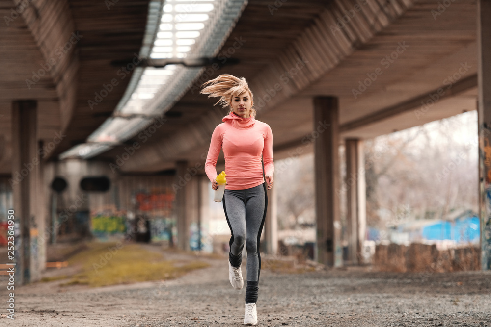 Beautiful blonde woman dressed in sportswear running under the bridge and holding bottle with water.