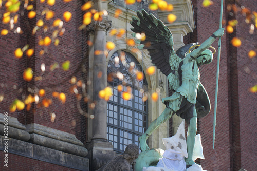 Ancient antique green metal Fontaine Saint Michael statue with yellow orange flower bogae in front of christian church at Hamburg Germany