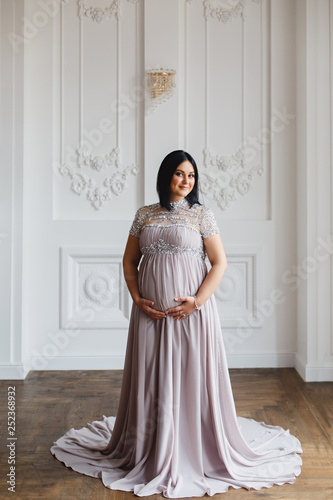 Young pregnant woman in a light classic interior. Caucasian woman with black hairs in long romantic dress near the large window. Concept of new life. Waiting of baby. Pretty pregnant woman in a room