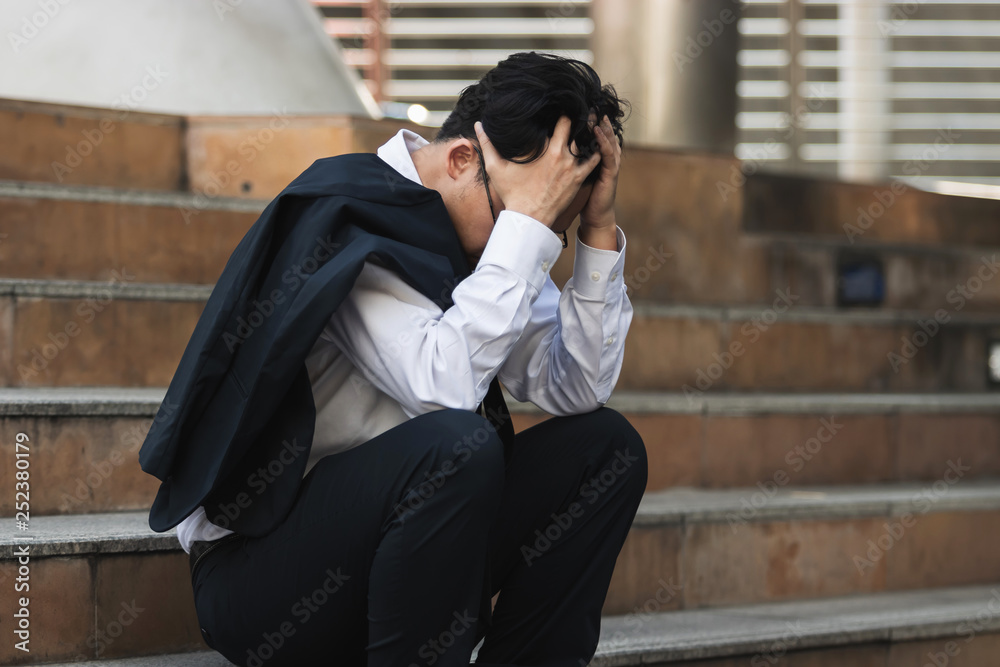 Upset stressed young Asian business man in suit with hands on head sitting on stairs. Unemployment and layoff concept.