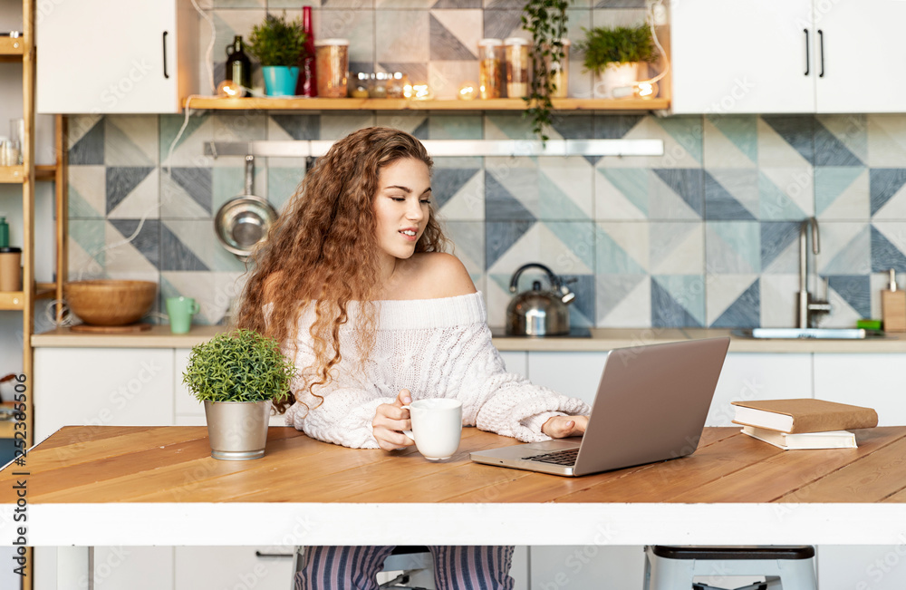 Portrait of young blogger girl using modern laptop and wearing comfy knitted white sweater. Sweet curly lady holding cup of tea. Technology and nice interior concept Portrait of young blogger girl using modern laptop and wearing comfy knitted white sweater. Sweet curly lady holding cup of tea. Technology and nice interior concept