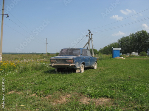 Coche antiguo en un parado ruso. Fondo hierba verde y arboles y azules manches de coche  casita y cielo