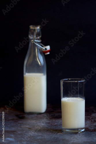 Full glass bottle of milk beside tall cup on wooden surface with copy space over dark background SELECTIVE FOCUS