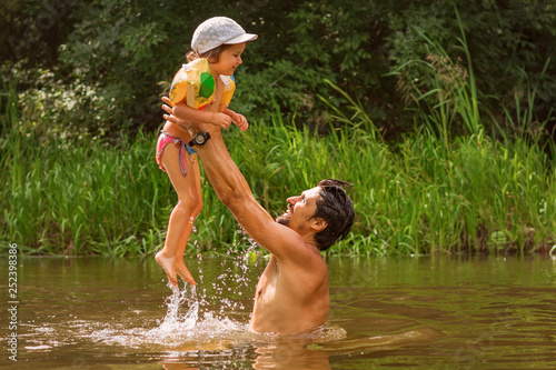 Fototapeta Naklejka Na Ścianę i Meble -  Dad and little daughter have fun on the river, the male throws the girl up, baby in inflatable sleeves