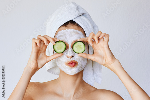 Beautiful young woman with facial mask on her face holding slices of cucumber. Skin care and treatment, spa, natural beauty and cosmetology concept.