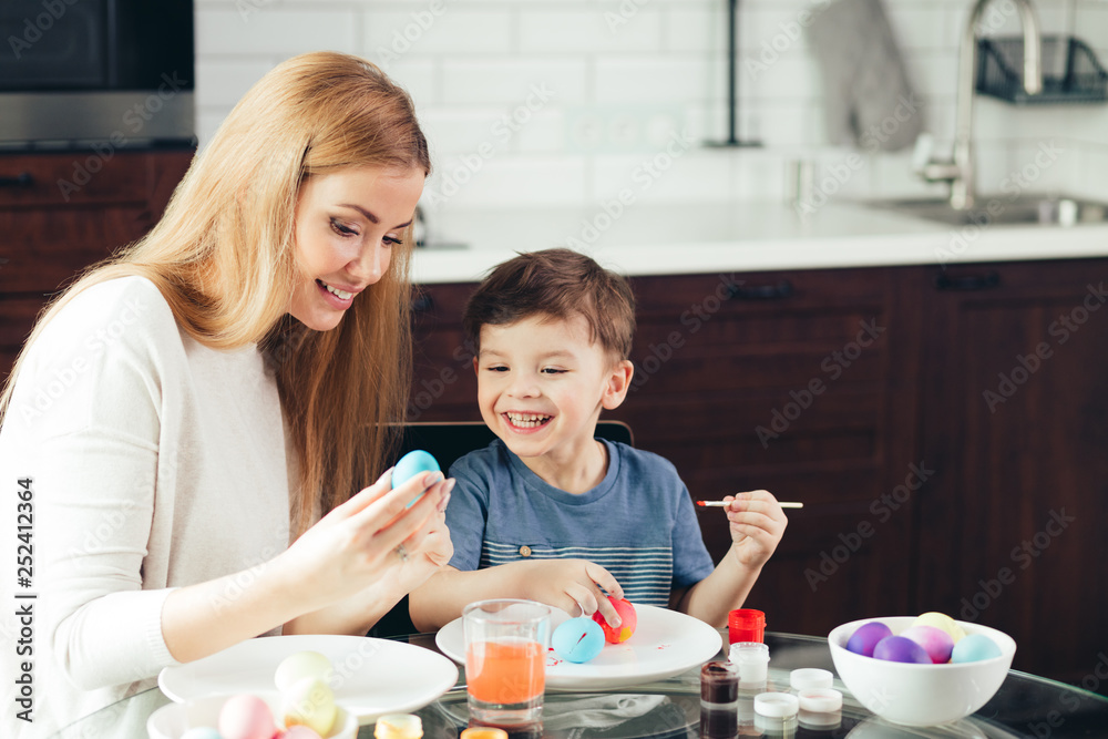 Blonde Female babysitter having confidence, skill and understanding to support children's play, interacting with boy in a playful way, painting eggs to Easter celebration.