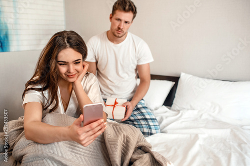 Young couple in the bed. Smiling indifferent woman with her phone is ignoring her unhappy man with a gift