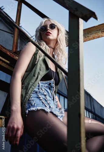 Beautiful young grunge (rock) girl in shorts, ripped pantyhose, shirt, vest and black glasses sitting on ladder. Outdoor portrait of informal model.