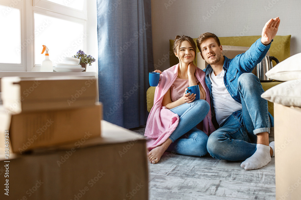 Young family couple bought or rented their first small apartment. People sit on floor and look up. Guy point with hand. Dreaming. Moving in. Young family couple bought or rented their first small apartment. People sit on floor and look up. Guy point with hand. Dreaming. Moving in.