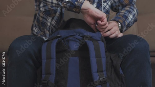 A man sits on the couch, puts things in a backpack, fastens it and leaves the frame.