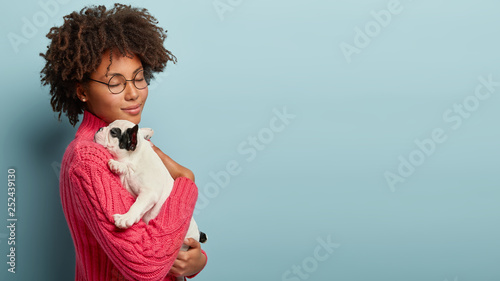 Beautiful dark skinned woman feels delighted, closes yes from pleasure, carries little baby dog with black and white colour, have fun at home, expresses tender emotions, care and love to small puppy