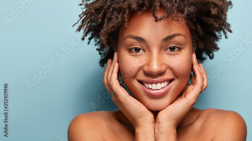 Close up portrait of relaxed black woman has gentle skin after taking shower, satisfied with new lotion, has no makeup, smiles tenderly, shows perfect teeth, stands shirtless against blue background