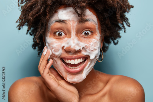 Pleased black lady has bubble mask, cleans facial skin with soap, has overjoyed expression, curly hairstyle, touches cheek, looks with pleasure, models over blue background, enjoys herself. Hygiene