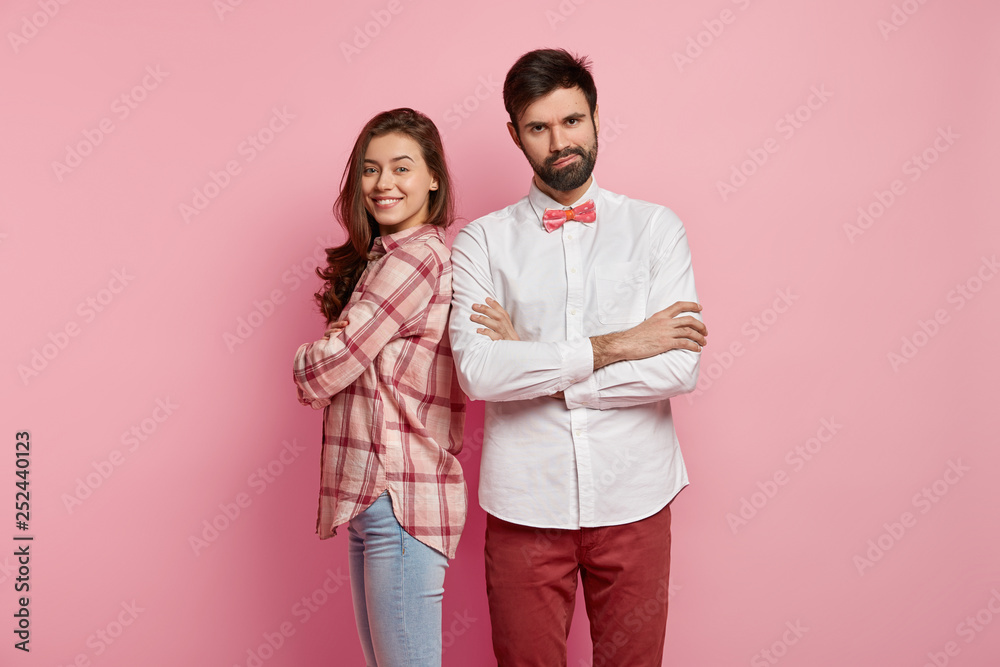 Upset angry young man with thick bristle, keeps arms folded, displeased with new assistant, wears elegant white formal shirt, happy female stands back to male partner, being sure in success. Upset angry young man with thick bristle, keeps arms folded, displeased with new assistant, wears elegant white formal shirt, happy female stands back to male partner, being sure in success.