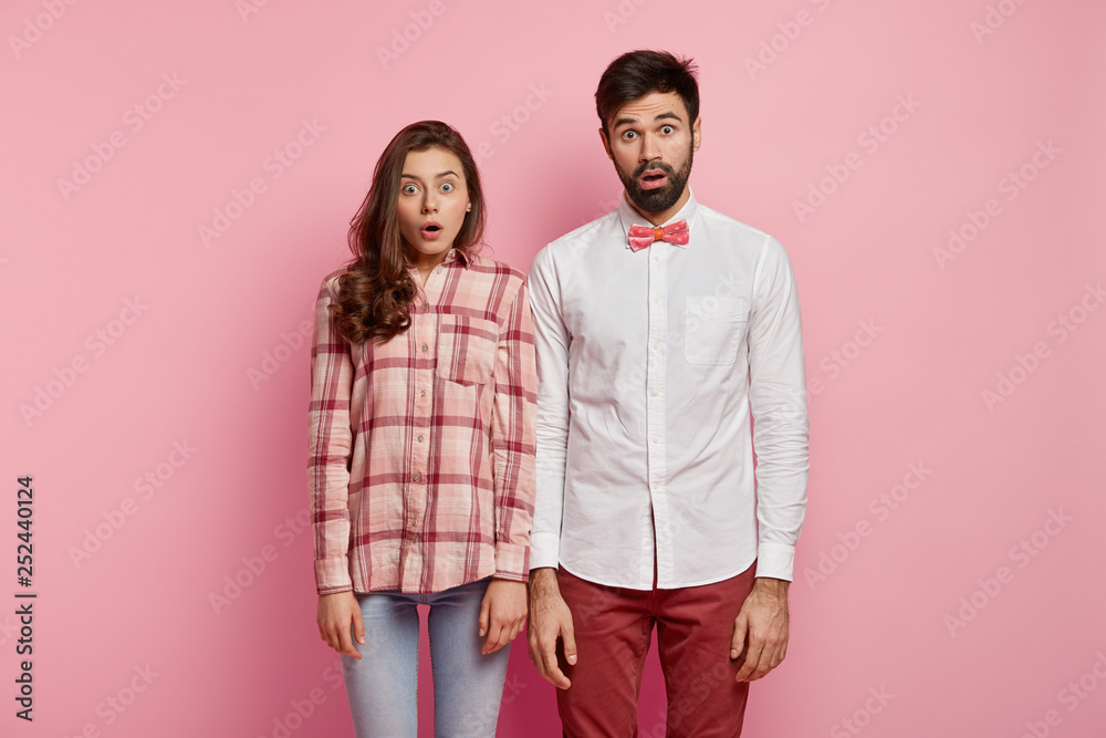 Studio shot of astonished woman and man have scared facial expressions, stare at camera speechless, feel stunned to hear shocking news, stand shoulder to shoulder, isolated over pink background Studio shot of astonished woman and man have scared facial expressions, stare at camera speechless, feel stunned to hear shocking news, stand shoulder to shoulder, isolated over pink background