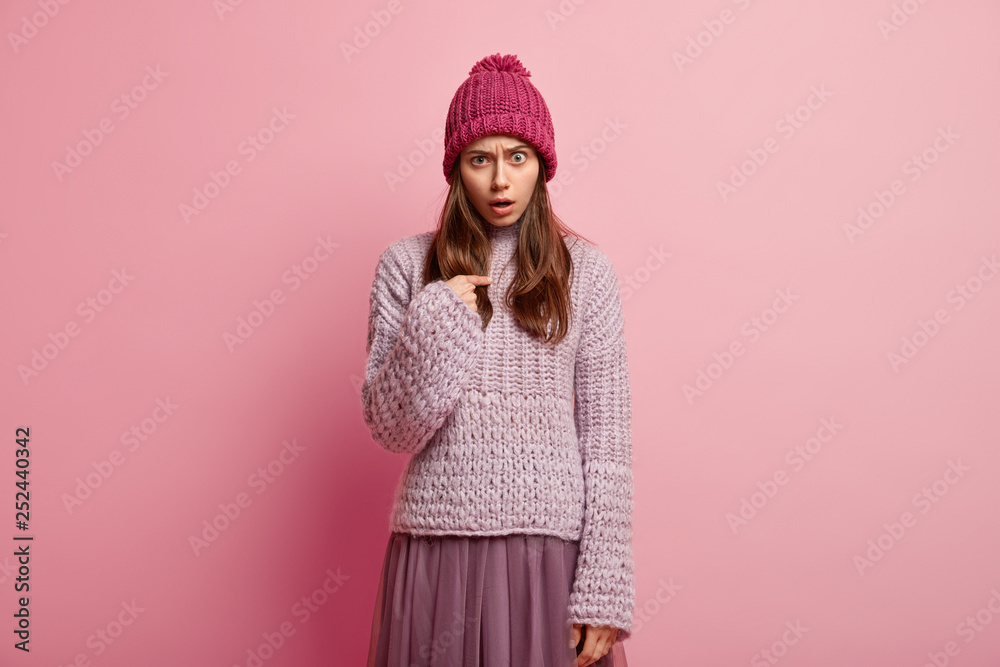 Displeased emotive European woman points at herself, wonders to loose game, looks with indignation at camera, wears casual winter clothing, models over pink background, has angry facial expression