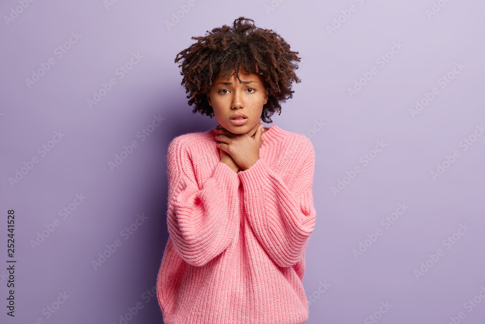 Upset discontent lady with dark skin, curly hair, keeps both hands on neck, suffers from asthmatic fit, wears oversized pink jumper, suffers from cough attack, poses indoor. Painful feelings in throat Upset discontent lady with dark skin, curly hair, keeps both hands on neck, suffers from asthmatic fit, wears oversized pink jumper, suffers from cough attack, poses indoor. Painful feelings in throat