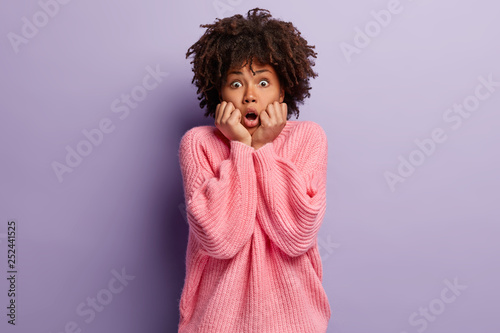 Neurotic African American woman looks stressfully at camera, keeps both hands near mouth, scared of fearful thing, dressed in pink oversized jumper, isolated over purple background. Omg concept