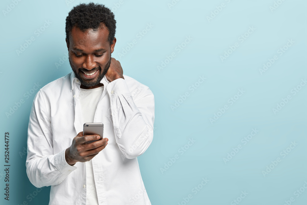 Smiling dark skinned male blogger creats new publication, holds modern cellphone, reads nice notification, sends money online, has bristle, isolated over blue background with mock up space aside Smiling dark skinned male blogger creats new publication, holds modern cellphone, reads nice notification, sends money online, has bristle, isolated over blue background with mock up space aside