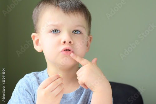 Emotional portrait of a white handsome caucasian blue eyed boy on neural blurred background. Cute sad toddler boy shows a finger on his mouth. Little boy with toothache