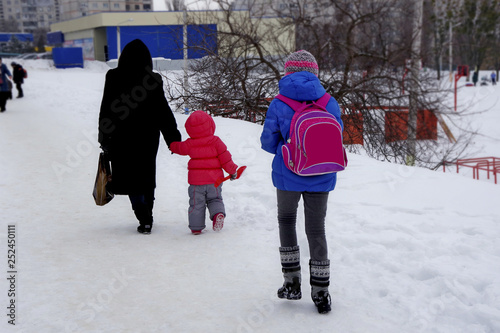 Winter in the city. Children are returning from school. A woman with a child and a girl with a backpack are walking along the sidewalk, which is covered with fluffy snow.