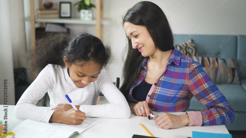 Adult mixed race woman teacher sitting and talking to american black student child in school class during day. Mother and daughter of different ethnicity are learning to draw together. Adult mixed race woman teacher sitting and talking to american black student child in school class during day. Mother and daughter of different ethnicity are learning to draw together.