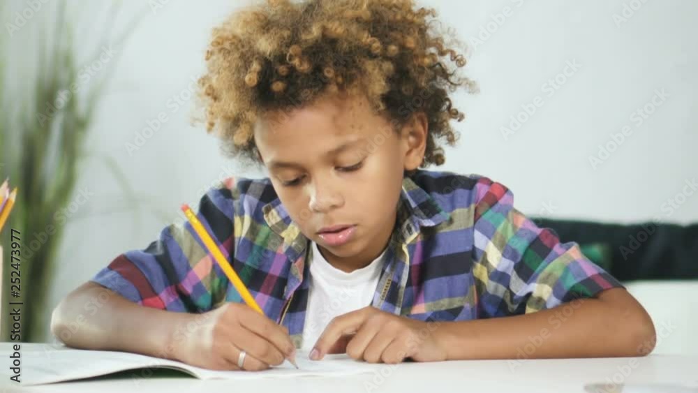 African american young boy is sitting at the dining table and doing school homework. Smart serious pupil is thinking, thoughtful and holding all attention on elementary lesson. Intelligent male African american young boy is sitting at the dining table and doing school homework. Smart serious pupil is thinking, thoughtful and holding all attention on elementary lesson. Intelligent male