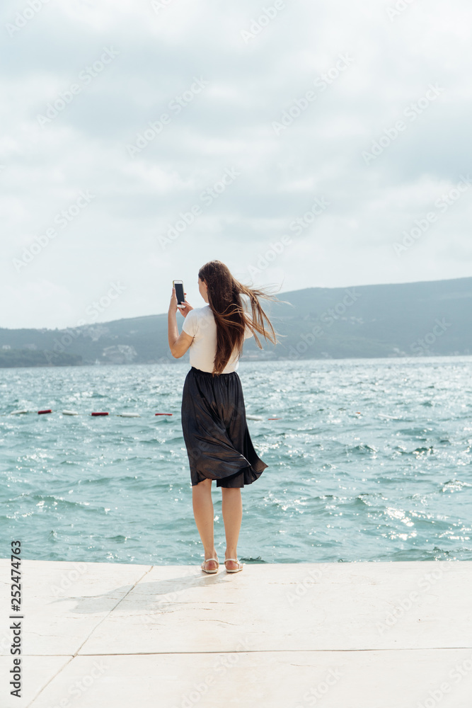 Young woman wear skirt use smartphone takingphotos of sea and mountains at stone pier. Summer vacation at european country. Girl travel on holiday. Woman enjoy and relax life. Summer vibes.