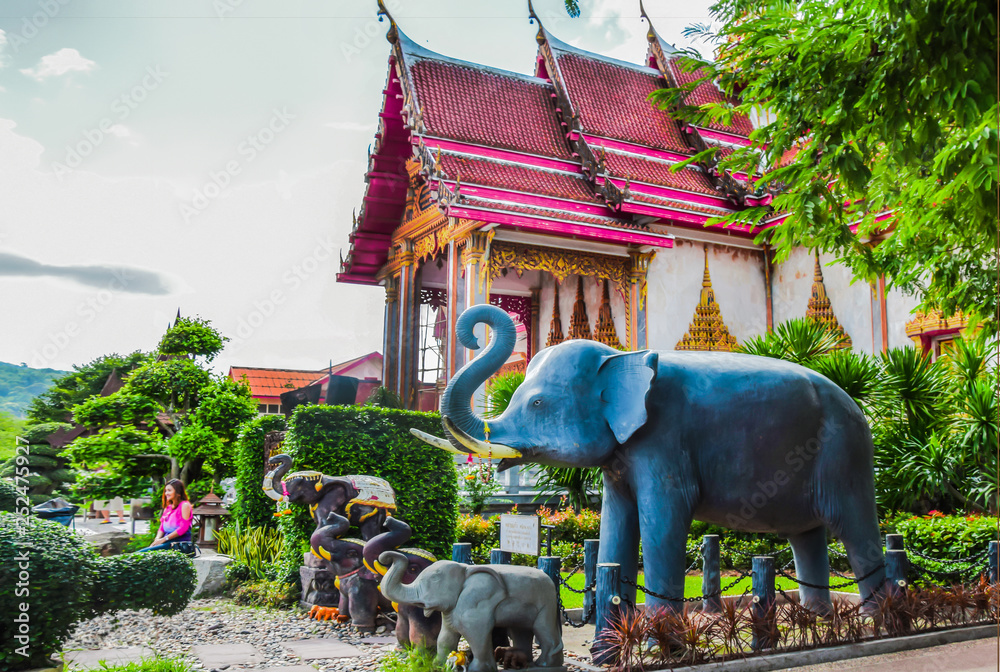 Elephant statues and elephants against the backdrop of a magnificent Buddhist temple among green trees on a sunny day. Thailand, Phuket Island