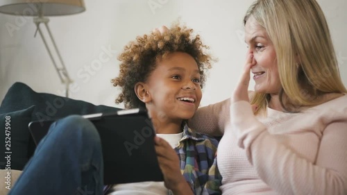 Glad and positive woman mom sitting on comfort couch inside bright light living room with her glad and excited son. They looking at tablet together, making wide beaming smile and clapping in palm