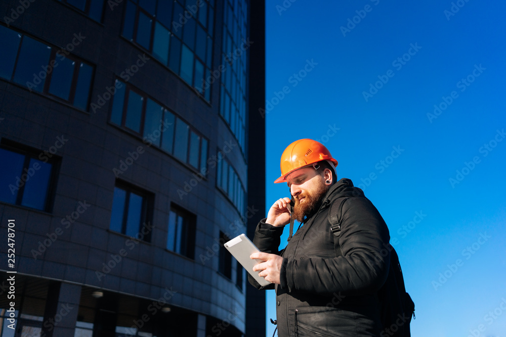 Pretty man - specialist civil engineer at orange hard hat is controls his project (outdoors). Blue sky and modern architecture (buildings) on background. Sunny day. Pretty man - specialist civil engineer at orange hard hat is controls his project (outdoors). Blue sky and modern architecture (buildings) on background. Sunny day.