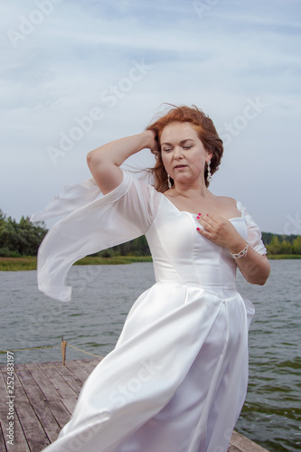 Red-haired bride in a white dress posing on a quay near a lake photo