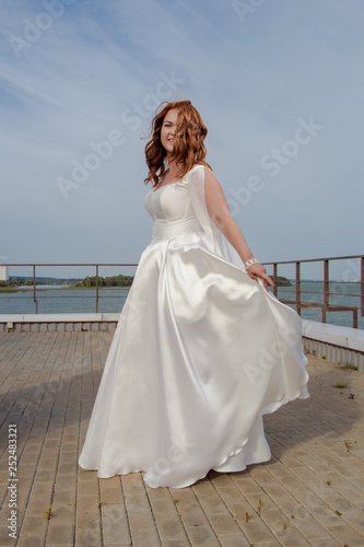 Red-haired bride in a white dress posing on a quay near a lake photo